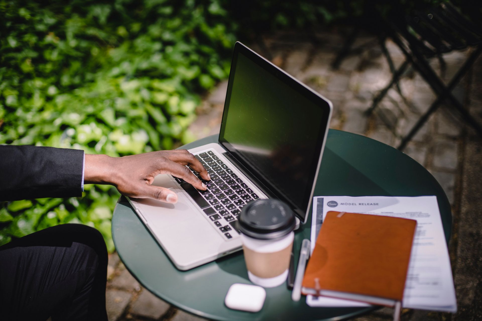 Business professional using a laptop outdoors with a coffee and notebook on a green table.