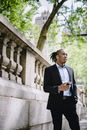 From below of serious young black male entrepreneur with dreadlocks in classy suit checking time on smartphone and waiting for meeting in city park