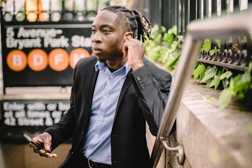 Free Confident young African American male in office suit listening to music on mobile through wireless earbuds while standing on stairs and looking away at subway entrance in summer Stock Photo