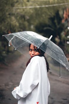 Woman in white holding a transparent umbrella, enjoying a rainy day outdoors. Aesthetic capture.