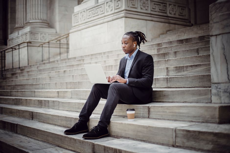 Full body serious African American businessman with earbuds wearing formal suit sitting on stone stairs of building and browsing modern netbook Full body serious African American businessman with earbuds wearing formal suit sitting on stone stairs of building and browsing modern netbook