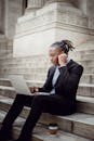 一位穿着西装的积极非裔美国商人坐在现代石制建筑的楼梯上，一边使用上网本工作，一边戴着无线耳机