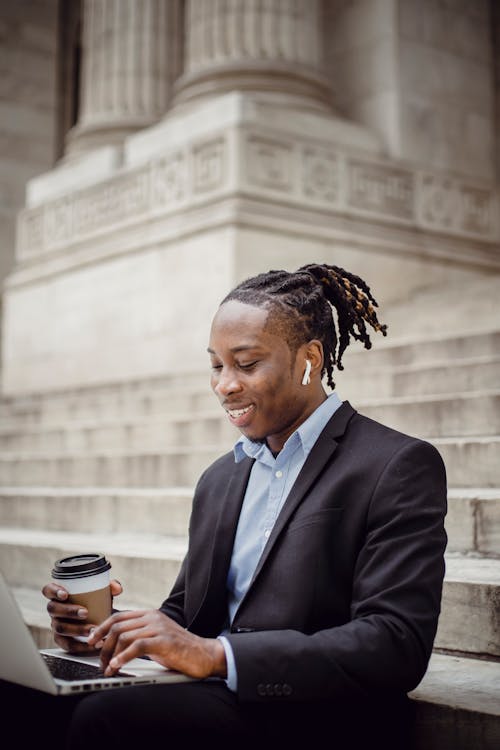 Free Cheerful black businessman typing on laptop on building stairs Stock Photo
