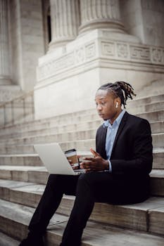 Businessman with earbuds working on a laptop outdoors on stone stairs, holding a coffee cup.