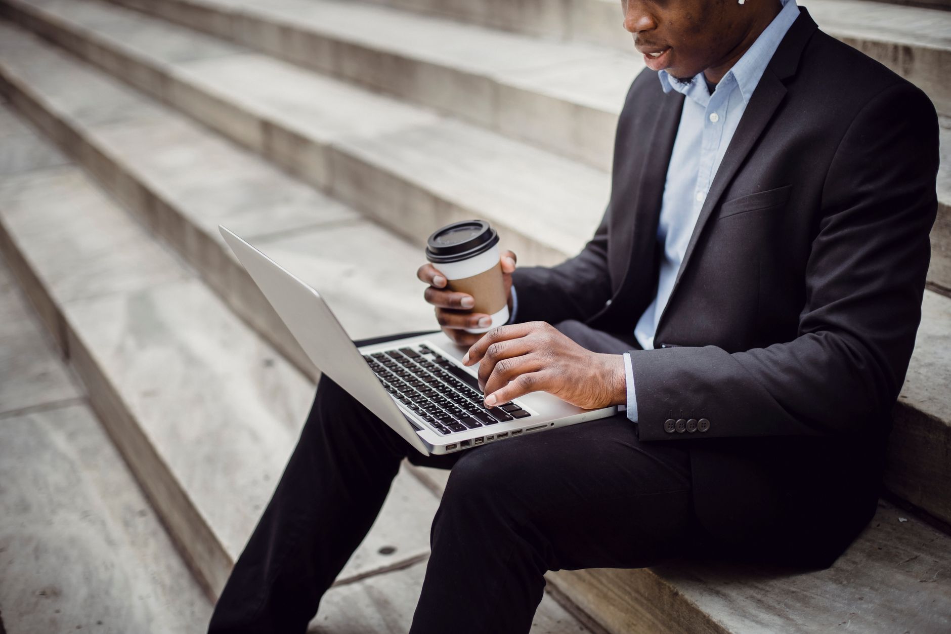Crop positive African American businessman wearing formal outfit sitting on stone stairs with paper cup of hot drink while working on project on laptop