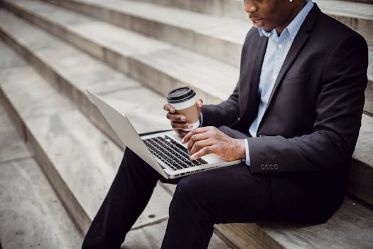 Crop positive African American businessman wearing formal outfit sitting on stone stairs with paper cup of hot drink while working on project on laptop