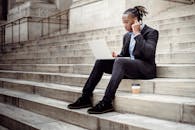 一位穿着正式黑色西装、神情专注的非裔美国商人，正坐在现代建筑的石楼梯上，戴着无线耳机浏览上网本