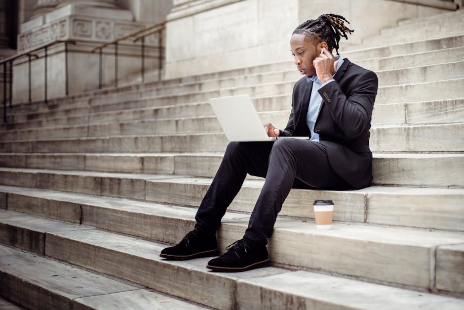 Full length focused African American businessman in formal black suit sitting on stone staircase of modern building and browsing netbook while putting on wireless earbuds