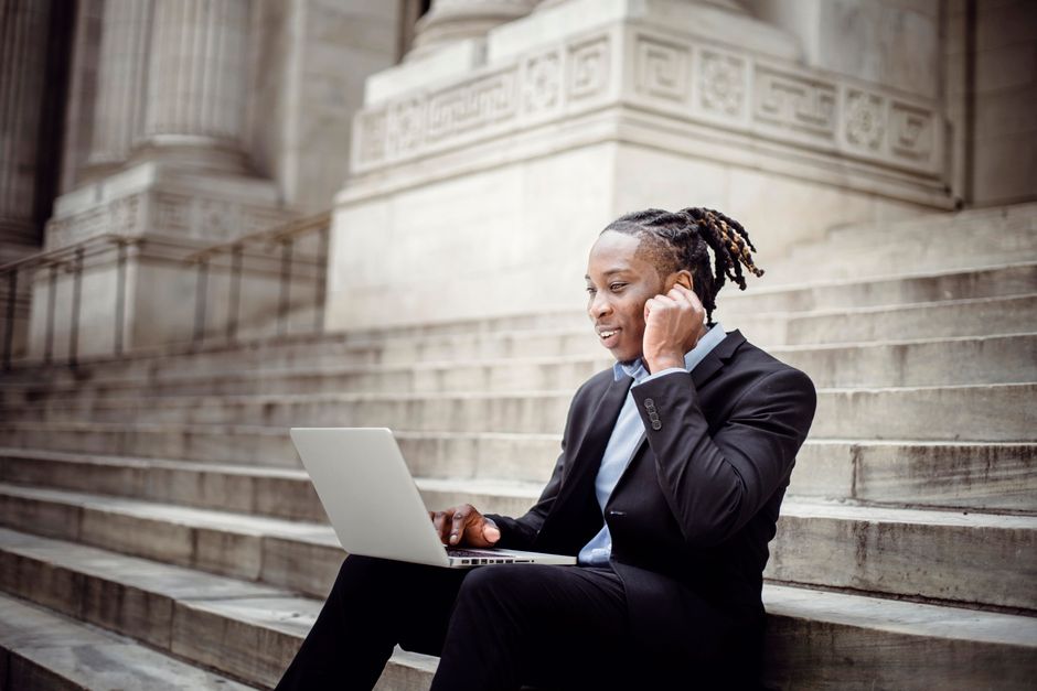 Positive smiling African American businessman in formal black wear answering video call via modern netbook and earbuds while working remotely on building stairs