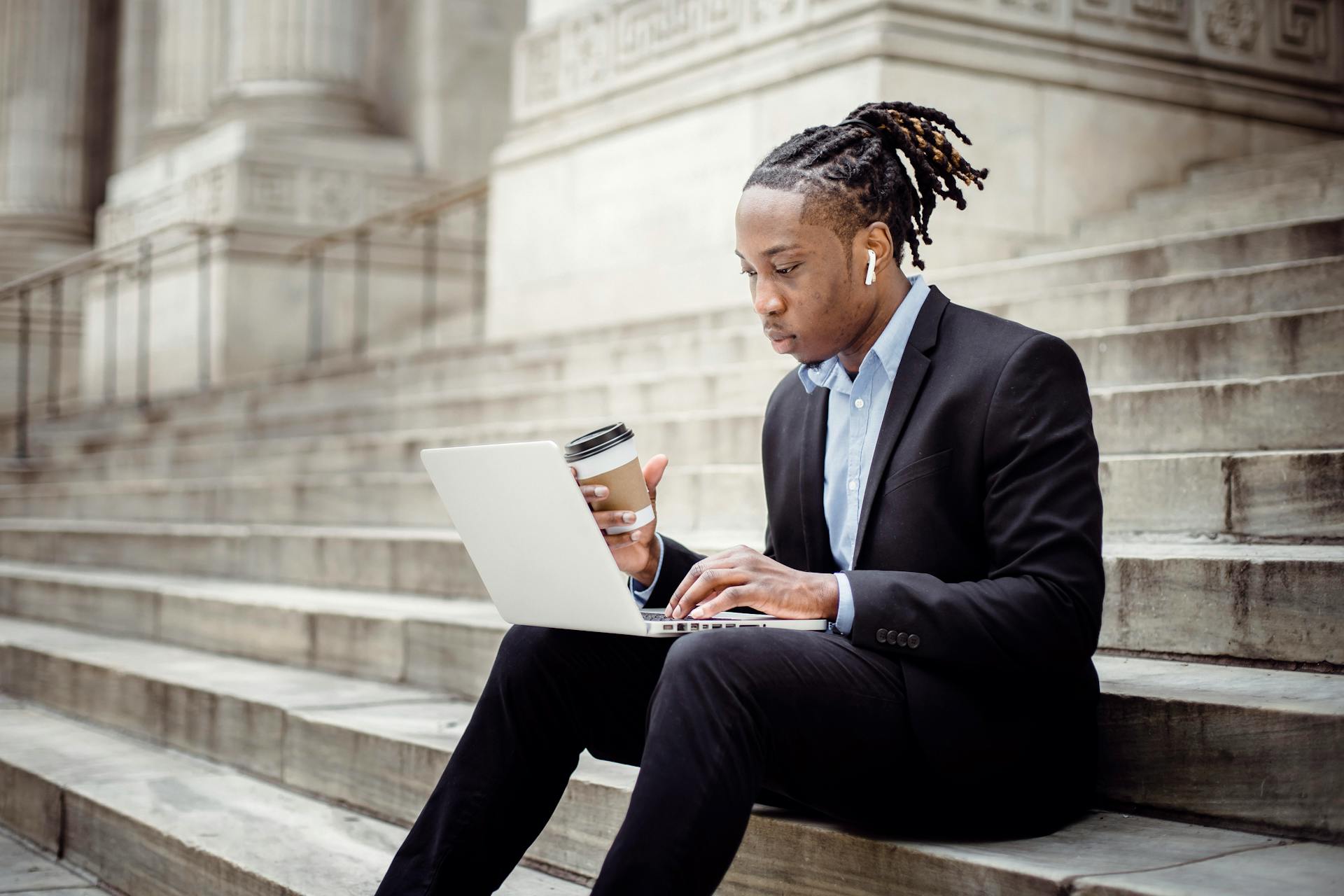 Concentrated African American businessman wearing formal clothes with coffee to go and earbuds browsing netbook while working remotely on stone building stairs