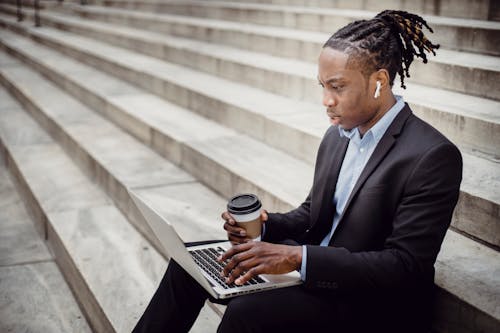 Concentrated perplexed black businessman wearing formal clothing sitting on street stone stairs with earbuds and coffee to go while typing on netbook keyboard