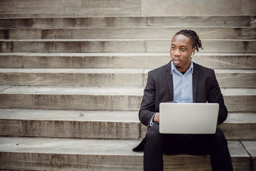 Positive young black businessman using laptop on stairs
