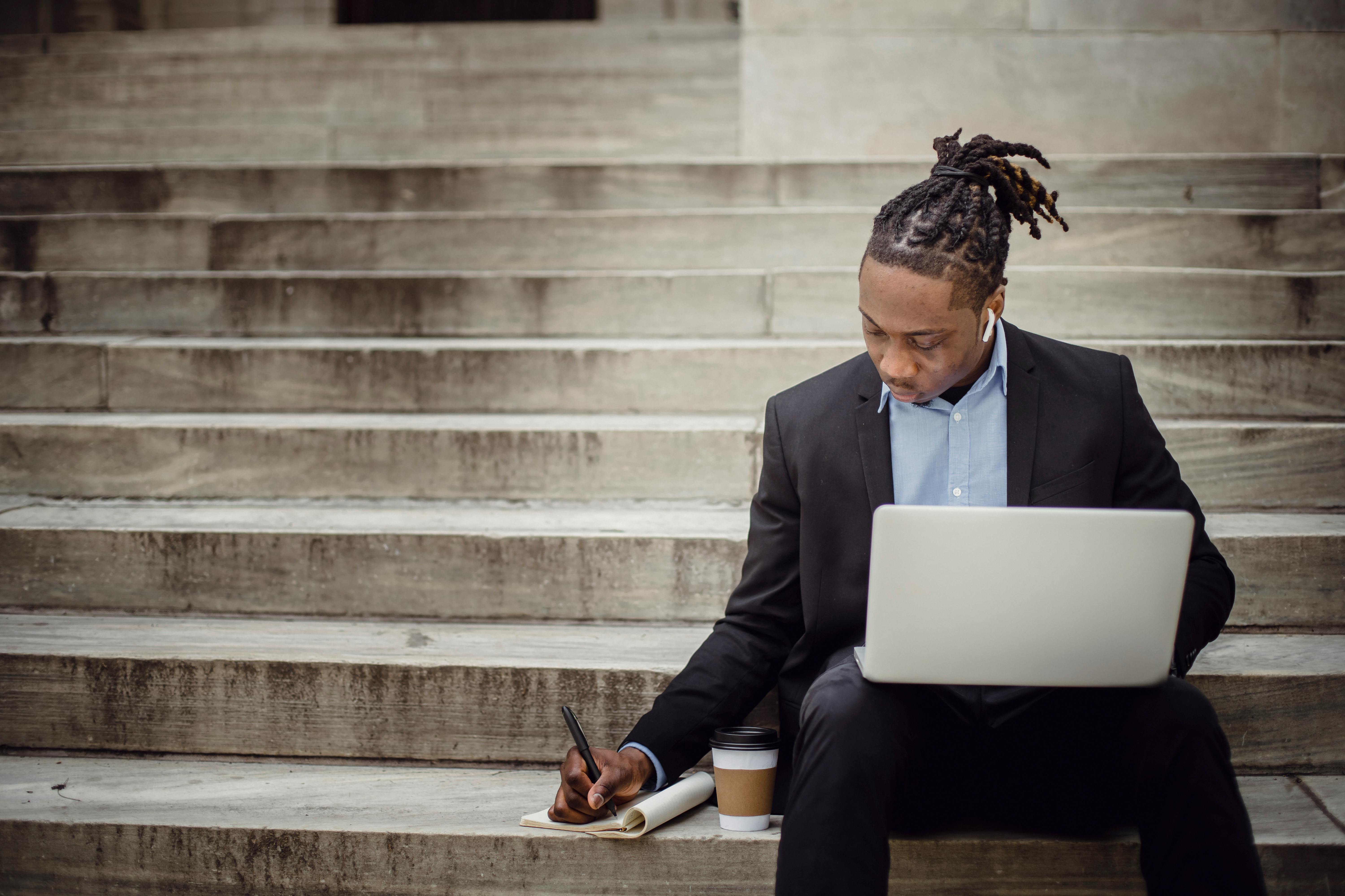 serious ethnic male manager taking notes while working on netbook on steps