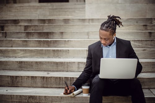 Concentrated young black man with dreadlocks in elegant suit browsing laptop and writing out information in notebook while sitting on stairs with takeaway coffee