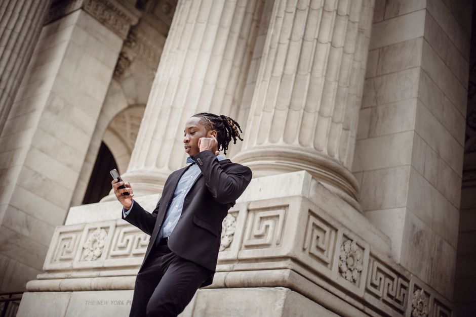 Low angle of serious well dressed black businessman with dreadlocks having phone conversation via wireless earphones while standing on stairs of columned building Low angle of serious well dressed black businessman with dreadlocks having phone conversation via wireless earphones while standing on stairs of columned building