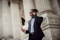 一位身穿正装、面带笑容的快乐年轻非裔美国男子，正站在现代建筑外通过手机进行视频通话