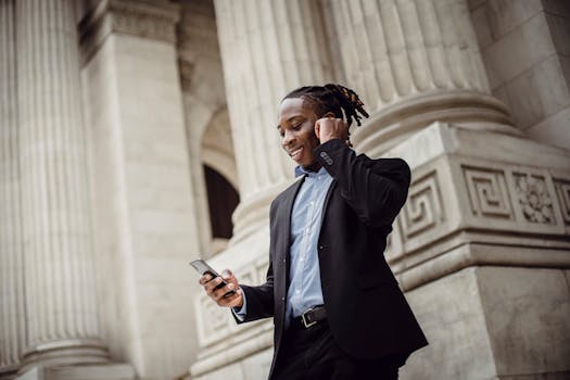 Cheerful young African American man in formal wear smiling and having video call via mobile phone while standing outside contemporary building