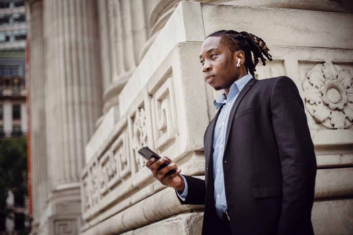 Free Pensive black businessman waiting on street with smartphone in hand Stock Photo