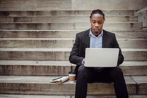 Focused young African American guy working on project using netbook while sitting on stairs with takeaway coffee and organizer