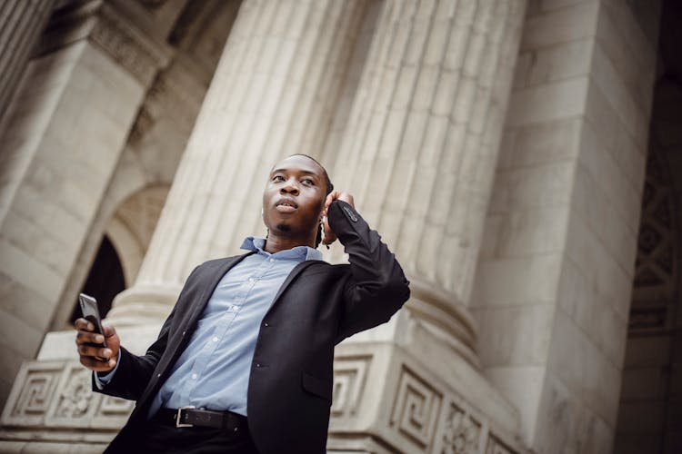 Thoughtful Black Male Executive Manager Talking On Smartphone Standing Near Pillars Of Building