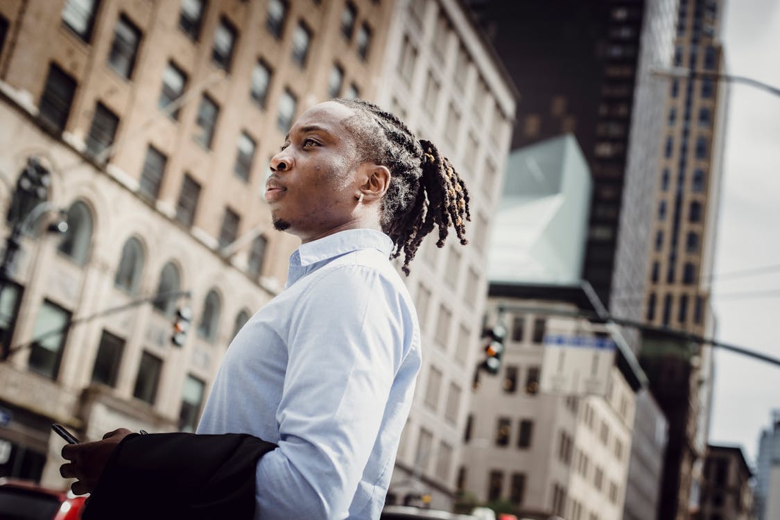 Free Side view of pensive young black man with dreadlocks in formal shirt standing on street in business district and looking away on sunny day Stock Photo