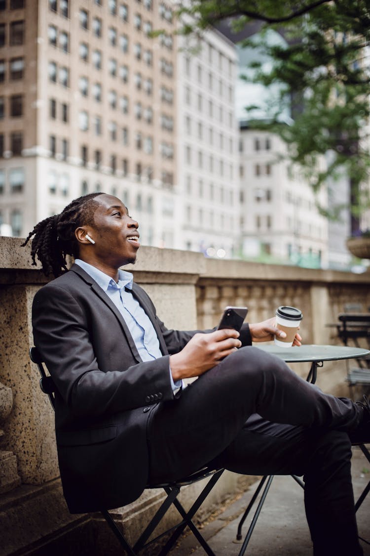 Cheerful Black Entrepreneur In Wireless Earphones Drinking Coffee On Terrace Of Business Center