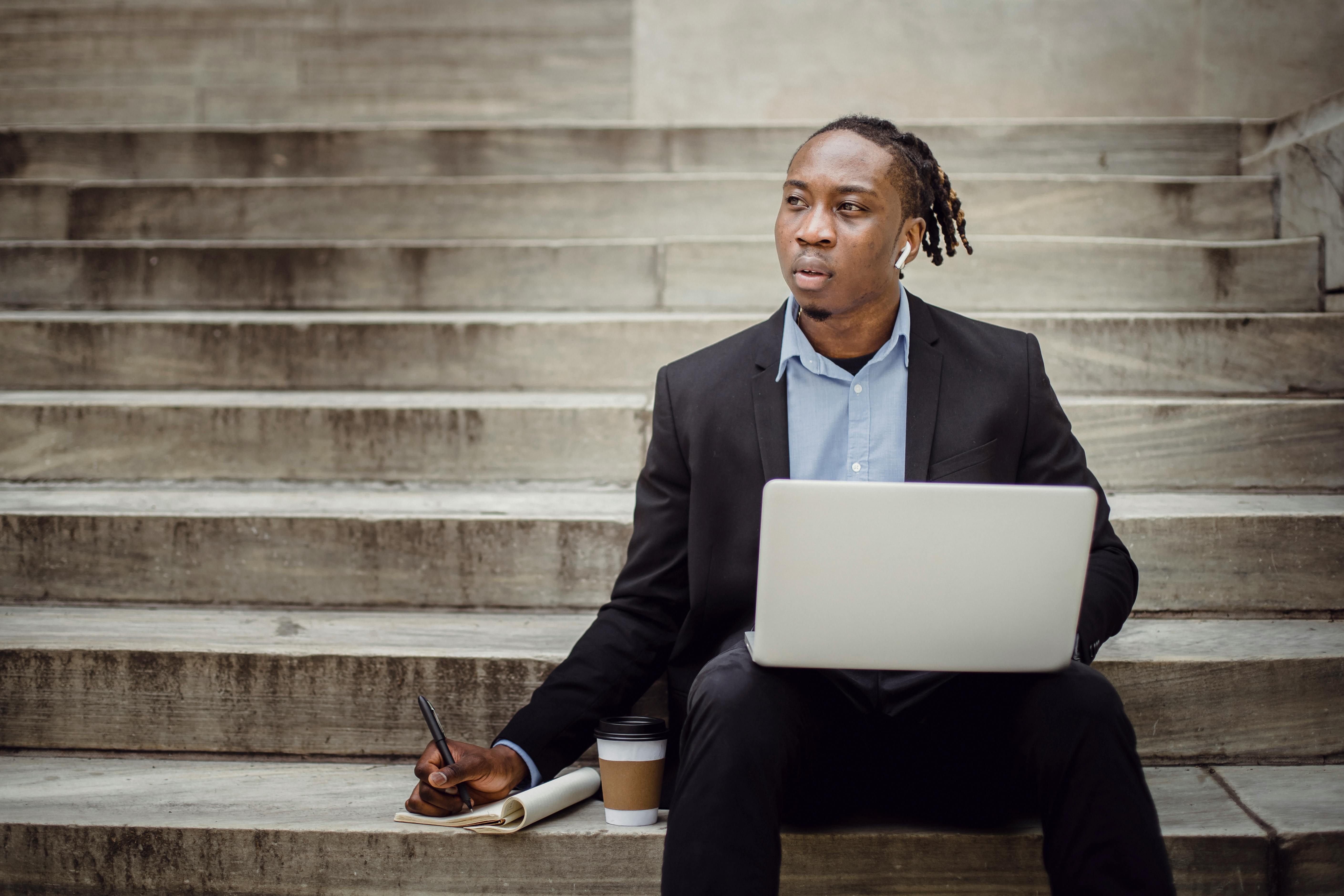 thoughtful black worker using netbook and taking notes sitting on steps