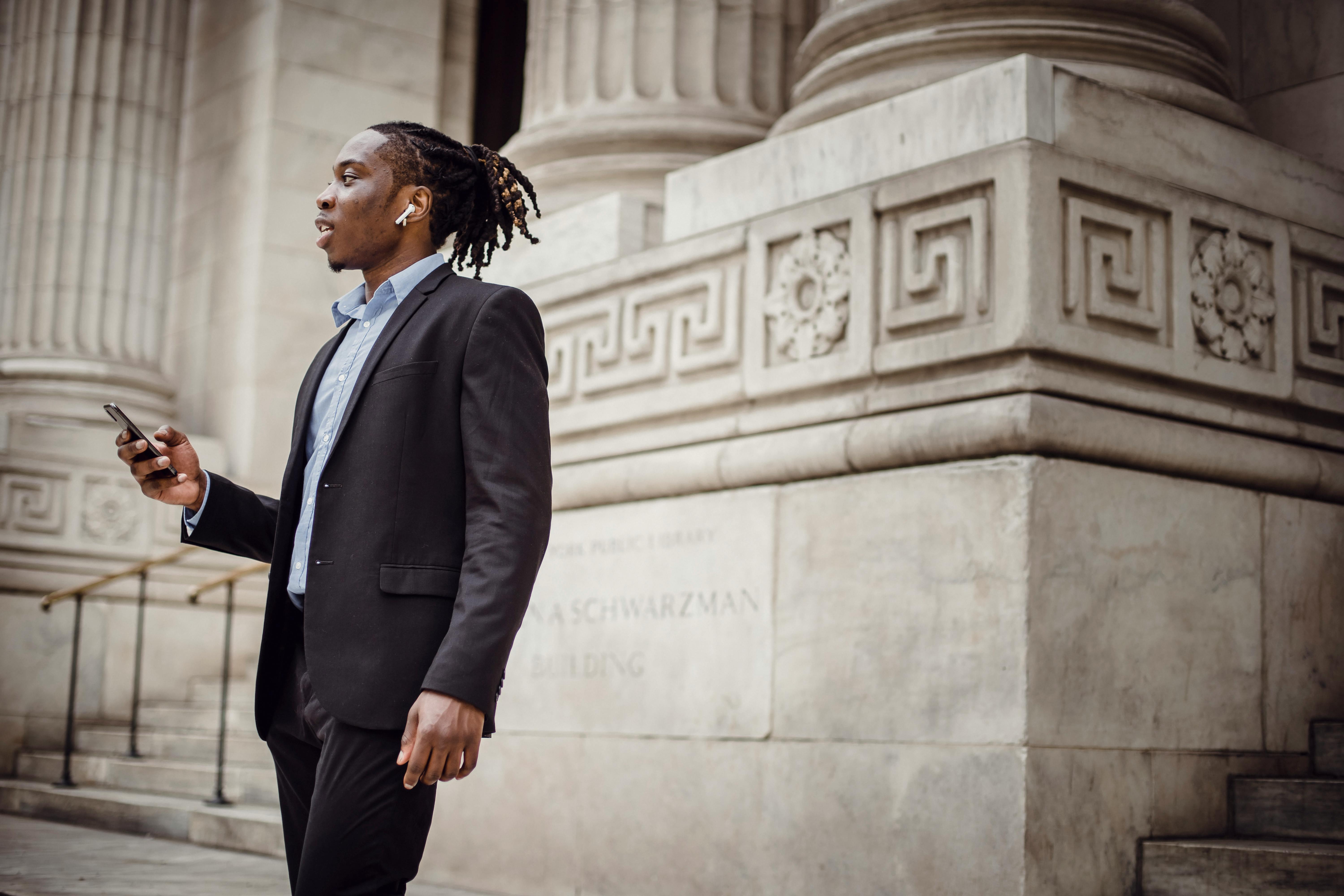 elegant black businessman using tws earphones while talking on smartphone against pillared building