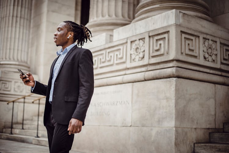 Elegant Black Businessman Using TWS Earphones While Talking On Smartphone Against Pillared Building