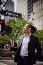 Young African American man in smart suit with dreadlocks and wireless earphones listening to music on smartphone while standing on street in Manhattan on sunny day