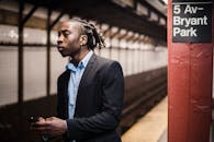 一名年轻而忧心忡忡的非裔美国男性通勤者，身穿正式西装，手持智能手机，在纽约地铁站等候列车