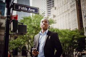 From below of young handsome black male in smart suit listening to music and waiting for car to pass while standing near street sign in Manhattan on sunny day