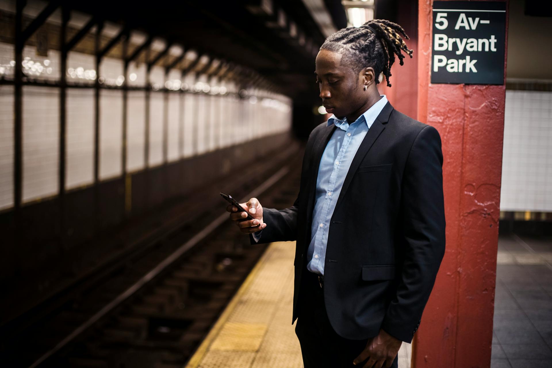 Modern young African American man in office suit text messaging on cellphone while waiting for train at Manhattan subway station