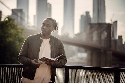 Young pensive African American male in casual clothes with open diary in hands leaning on balcony railing and looking away against blurred Brooklyn Bridge in summertime
