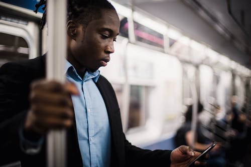 Young black male office employee in formal wear standing and using cellphone while commuting to work by train