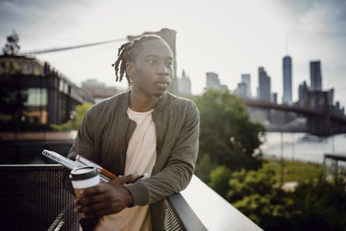 Dreamy African American student with dreadlocks in casual clothes standing leaned on balcony fence with plastic glass of coffee and organizer while contemplating river and looking away in morning