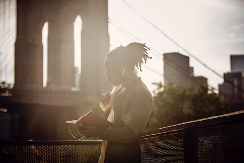 Side view of pensive young black man with braided hairstyle reading book and listening to music with wireless earphones while standing on city promenade against bridge