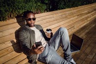 从上方看去，一位身穿时尚服装、戴着太阳镜的年轻黑人男性正坐在公园的木凳上，一边喝着咖啡，一边用笔记本电脑，还拿着手机自拍