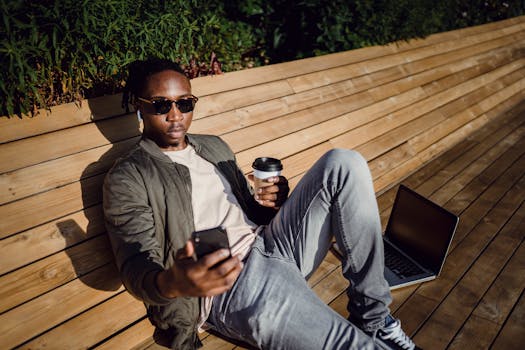 从上方看去，一位身穿时尚服装、戴着太阳镜的年轻黑人男性正坐在公园的木凳上，一边喝着咖啡，一边用笔记本电脑，还拿着手机自拍