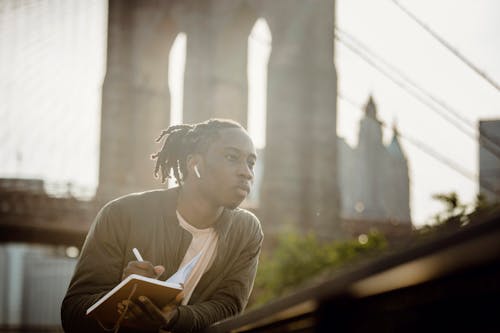 Thoughtful young black man with dreadlocks listening to audio story in wireless earphones and writing down information in notebook while leaning on fence on city promenade