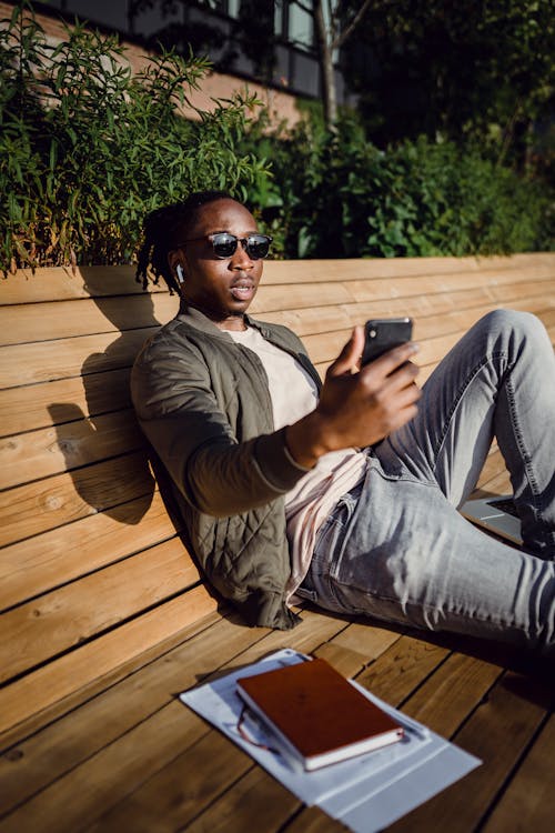 Young black guy chatting on smartphone sitting on wooden bench