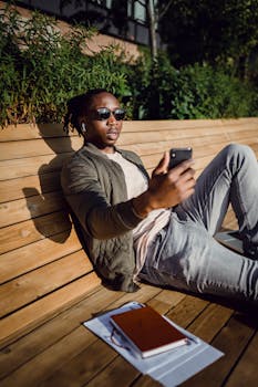 Casual young man enjoying a sunny day on a bench while using his smartphone. Perfect for lifestyle and tech themes.