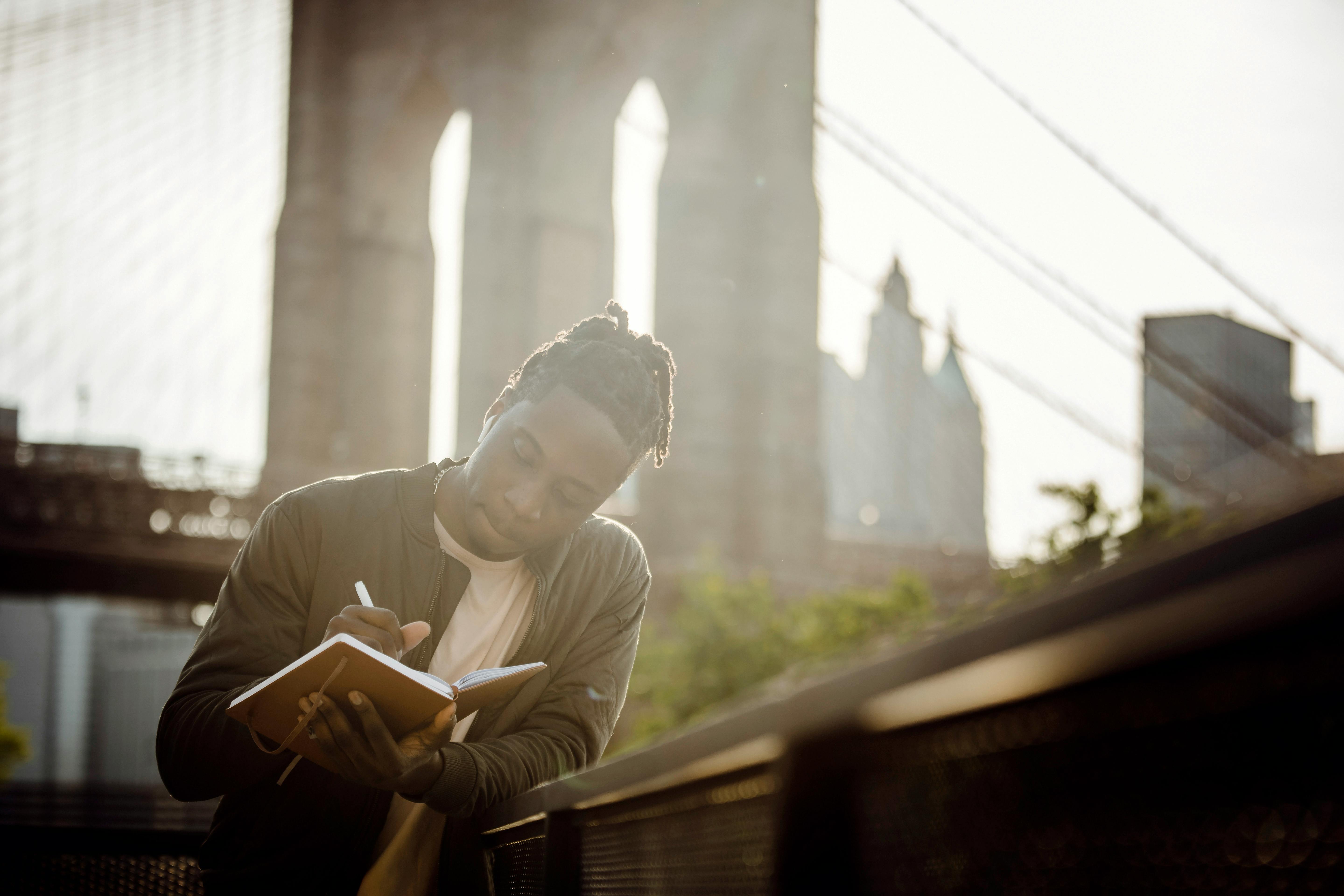 Low angle of concentrated young African American male journalist writing thoughts in notepad while chilling on city riverbank on sunny day