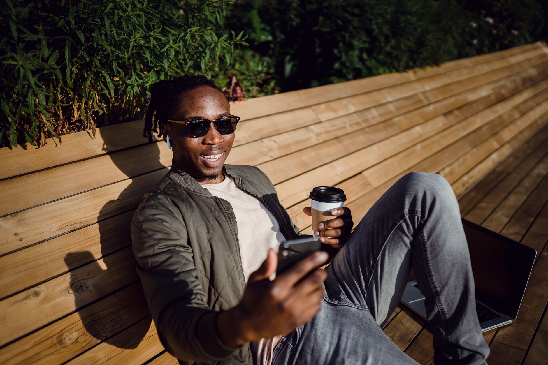 From above of happy African American guy in casual wear and sunglasses having video chat on smartphone while chilling on bench in park with takeaway coffee