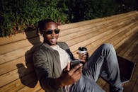 一个开朗的年轻人坐在公园的长凳上，喝着咖啡，拿着智能手机，享受着阳光明媚的一天