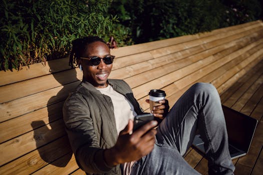 A cheerful young man enjoying a sunny day on a park bench with coffee and smartphone.