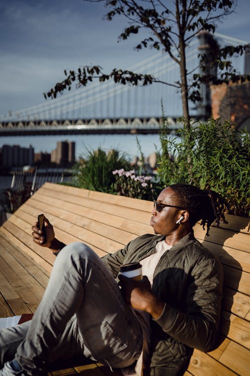 Focused black guy taking selfie on smartphone sitting on bench on embankment