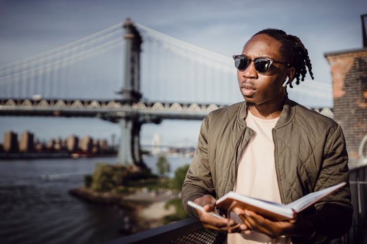 Young Black Man Standing On Embankment With Notebook In Hand