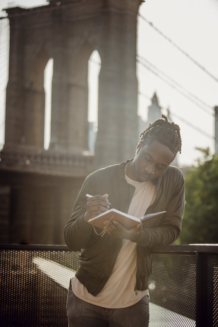 Pensive Black Man Taking Notes In Diary Against Bridge