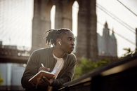 Positive black man listening to music with wireless earphones and taking notes in diary on street